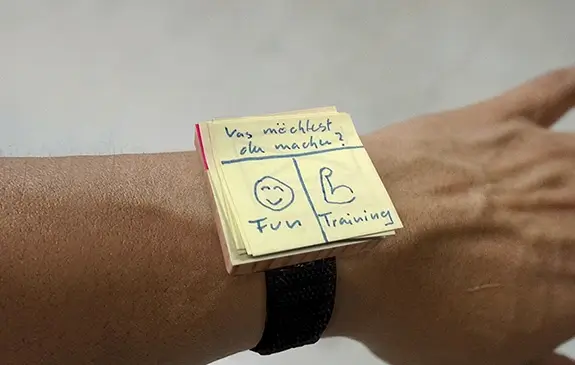 An arm whose wrist carries a low fidelity prototype of a smartwatch, made of a small wooden plate glued to a velcro fastener, carrying small postits and drawings of an app user interface, the headline 'What do you want to do today?', and the buttons 'Fun' and 'Training'