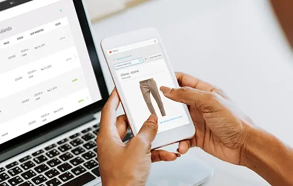 Looking over a person's shoulder on a smartphone, held in front of a notebook computer, both displaying an merchant's app user interface with fashion products to be managed